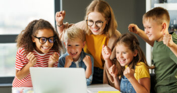 Lehrer und fröhliche Schüler blicken auf den Bildschirm ihres Laptops und feiern den erfolgreichen Abschluss einer gemeinsamen Schulaufgabe